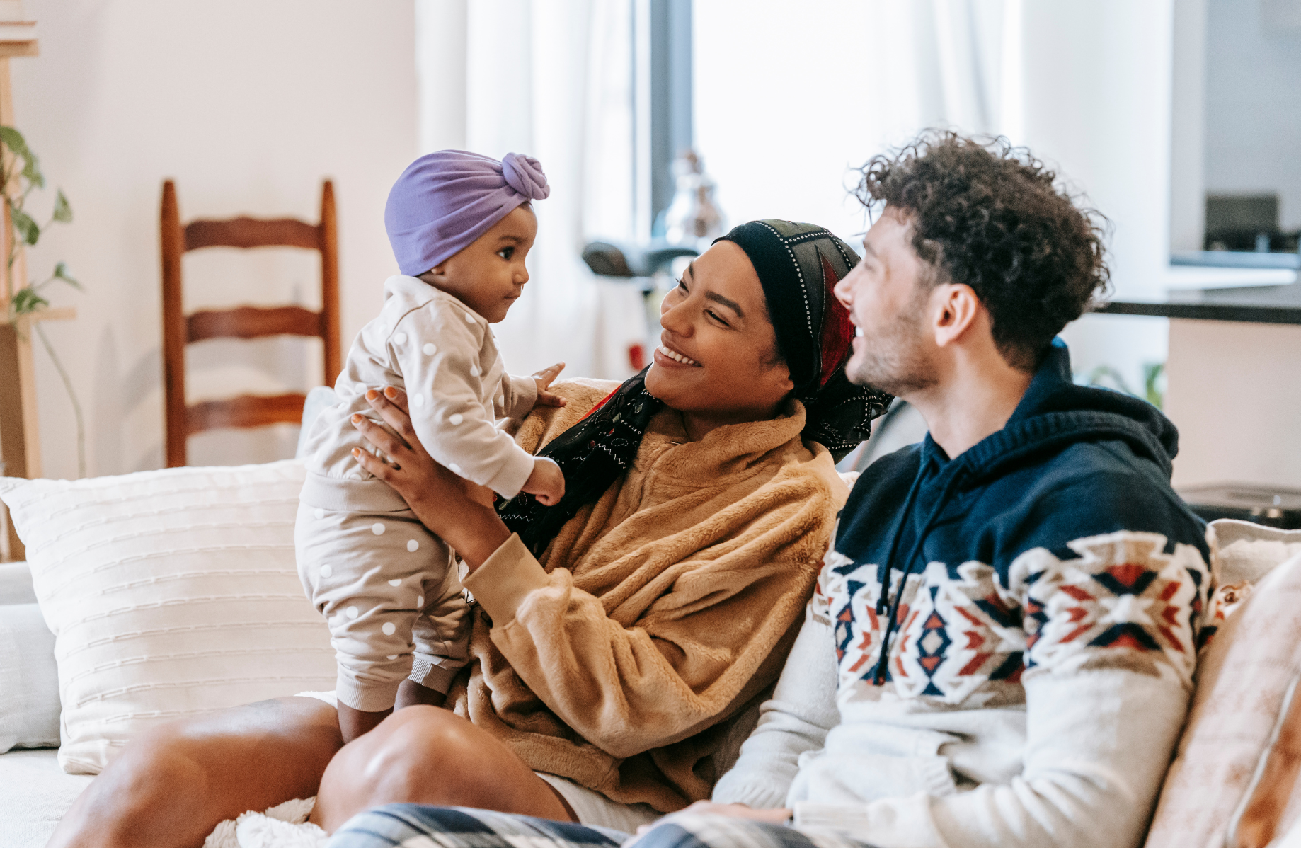 A young couple smiling at the toddler being held standing by one caregiver on their lap.